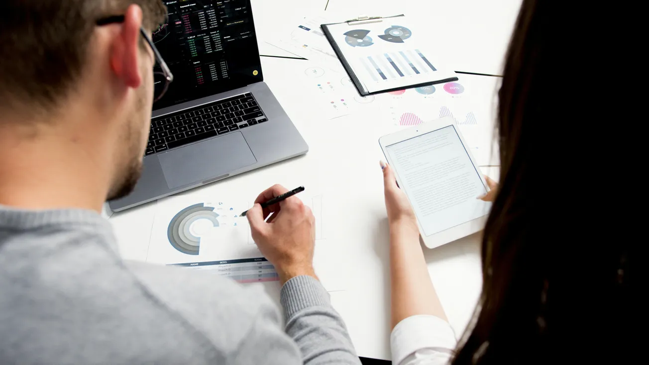 Two people collaborate at a desk with a laptop, analyzing data charts and graphs. One holds a pen, the other reads a tablet. 