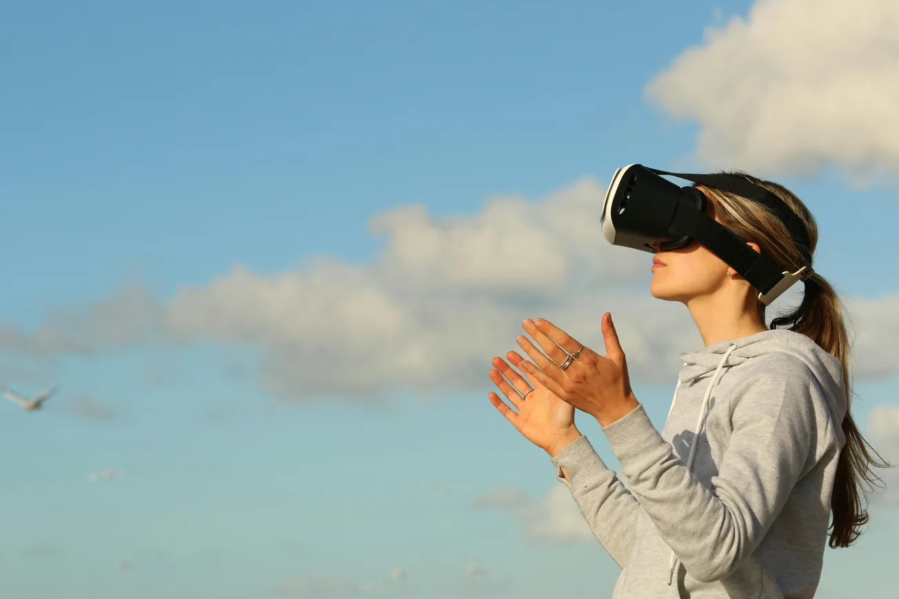 A person in a gray hoodie uses a VR headset outdoors, clapping hands against a bright blue sky with clouds. 