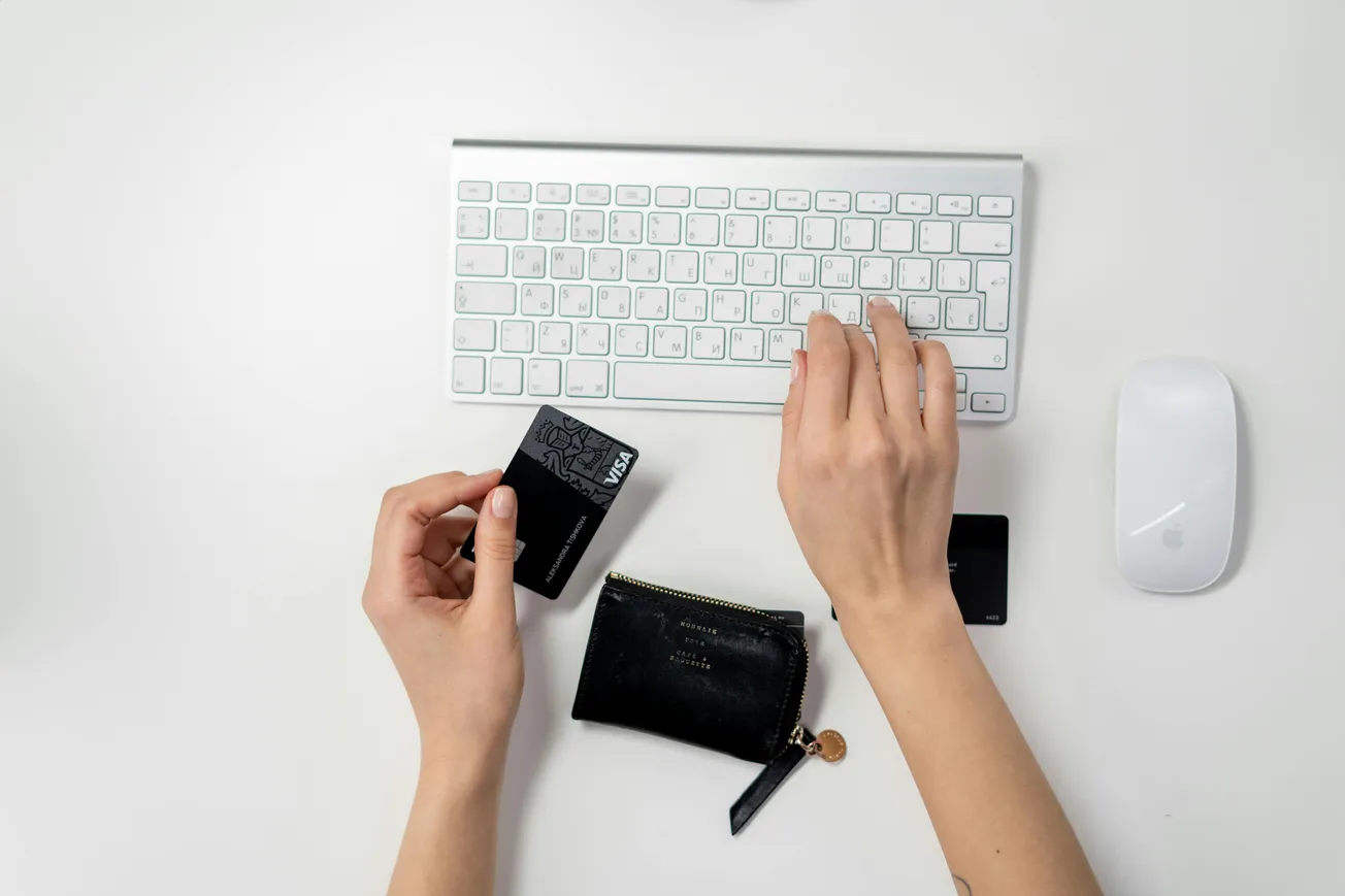 Hands using a computer keyboard and mouse, with a black credit card in one hand and a small black wallet nearby, indicating online shopping.