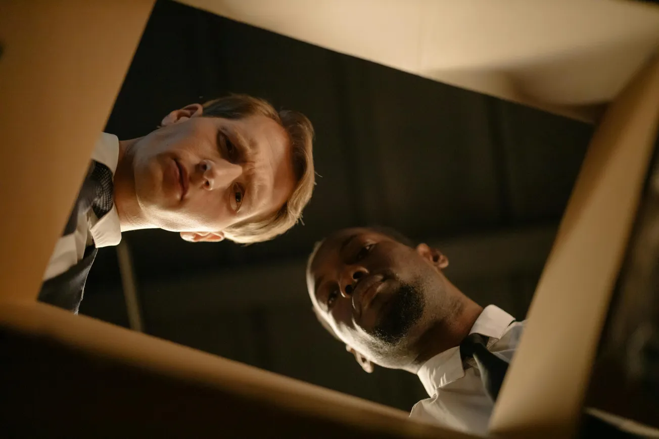 Two men in business attire looking down into a box with confused expressions. 