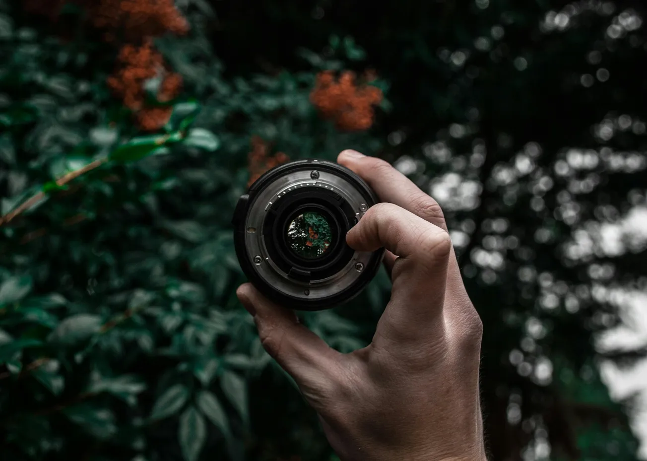 A hand holding a camera lens, framing a blurry background of lush green leaves and orange flowers.