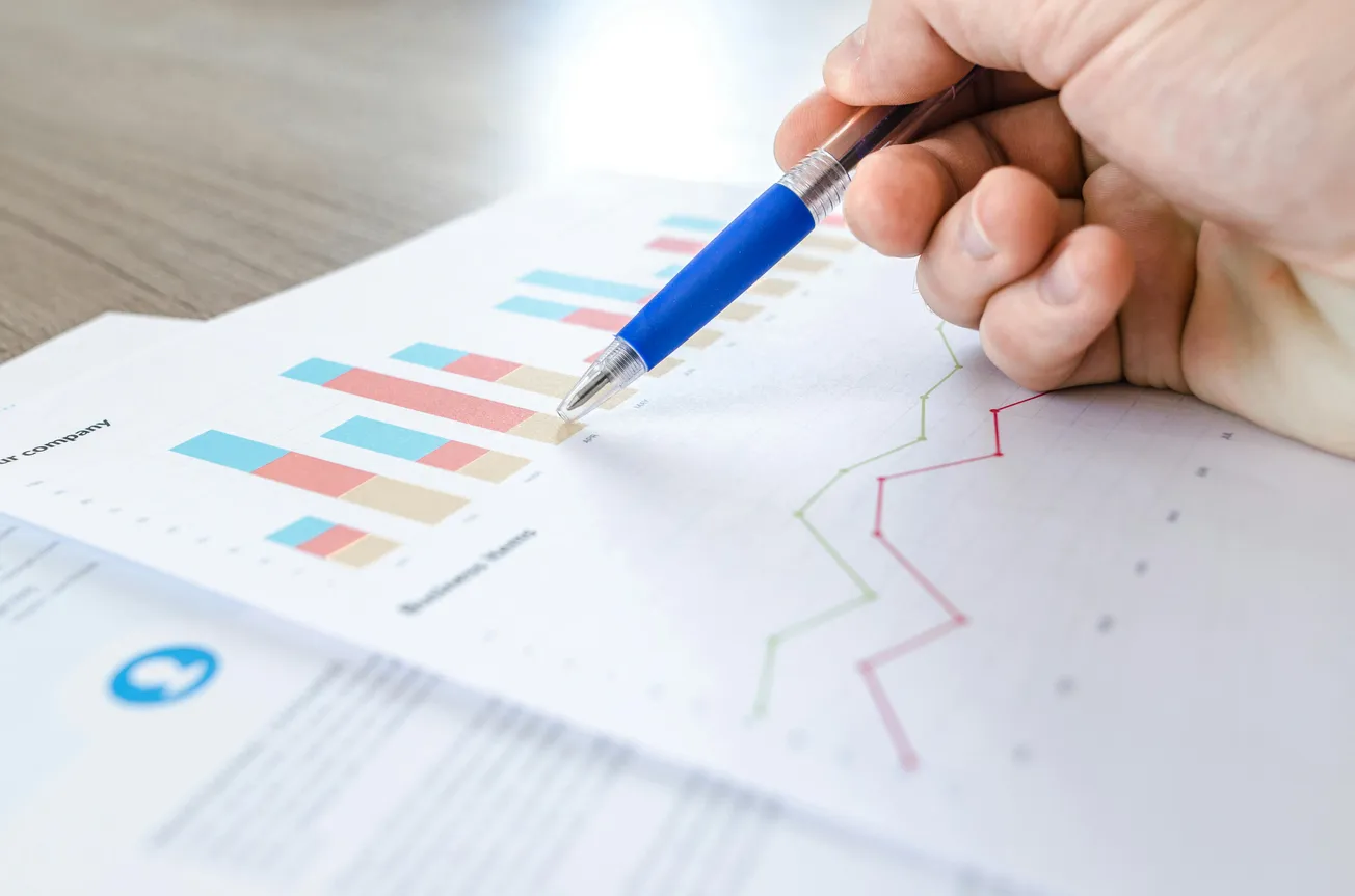 A hand holding a blue pen points at a line graph on a document, with a colorful bar chart visible in the background, indicating data analysis.