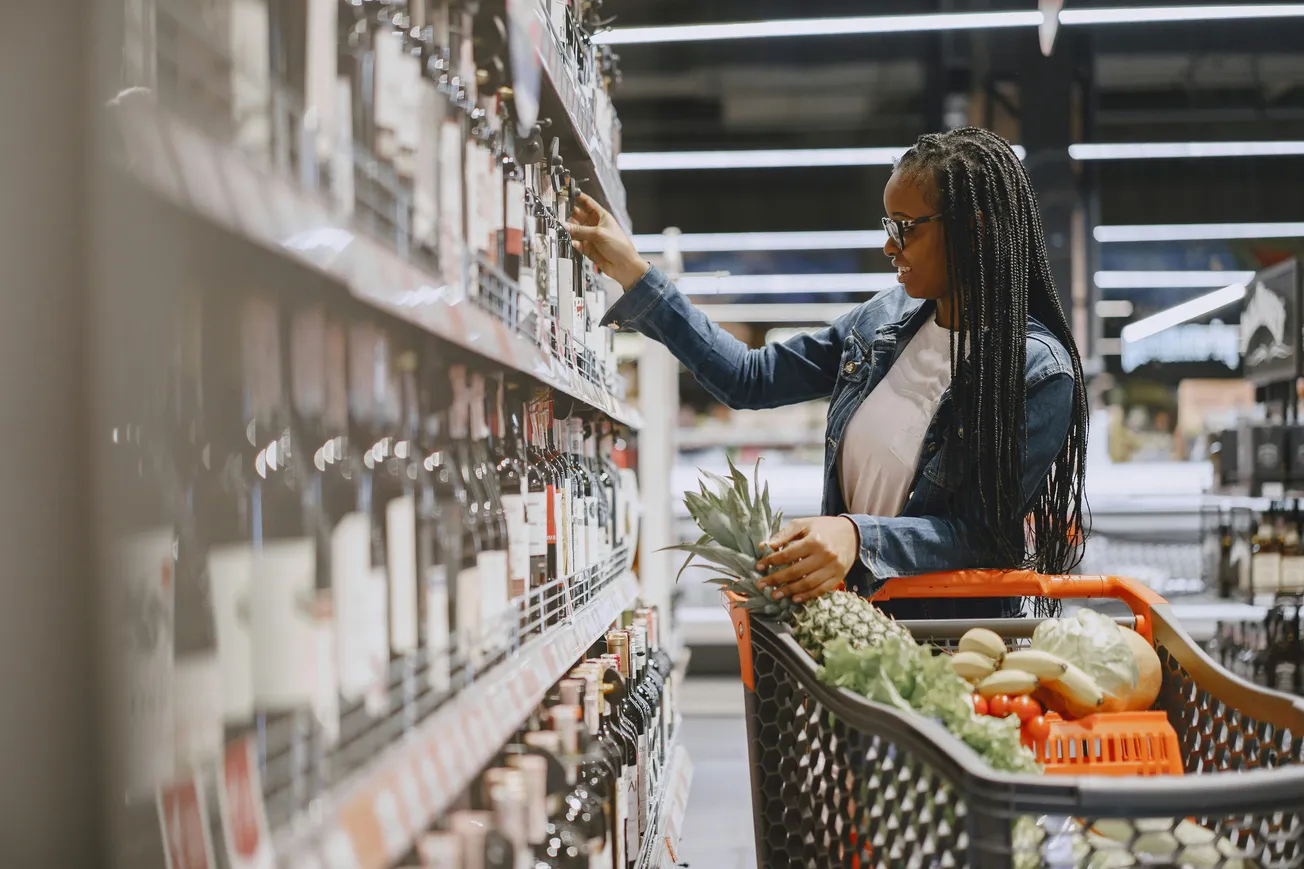 A young woman in a denim jacket shops in a grocery store, reaching for a wine bottle on a shelf. Her cart is filled with fresh produce.