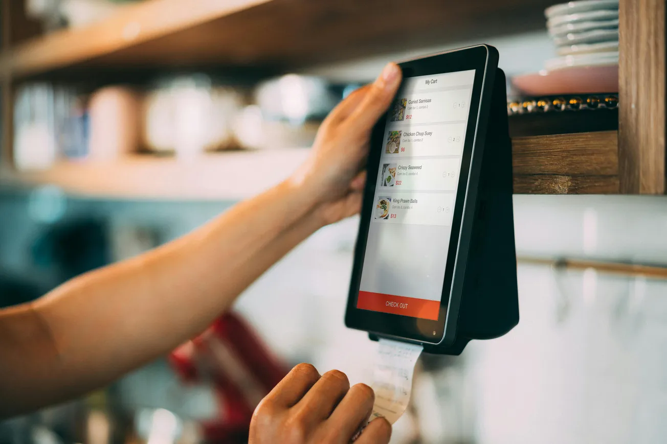 A person operates a touchscreen tablet mounted on a shelf, with an order list displayed. Below, a receipt is printing.
