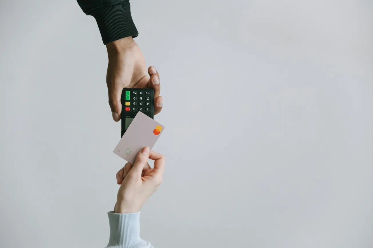 A hand holds a payment terminal while another hand brings a credit card toward it, tapping to pay.