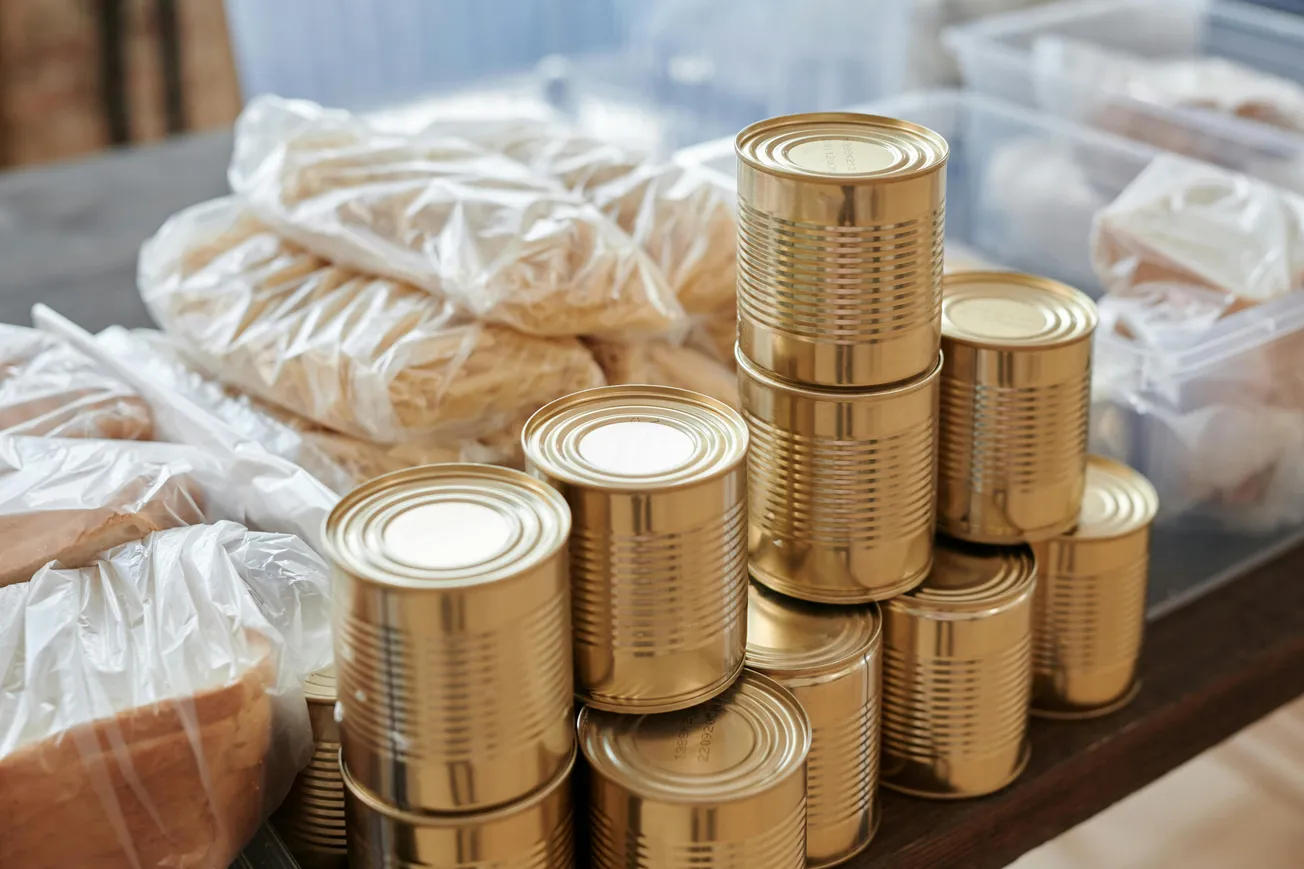 Stacked golden food tins and clear plastic bags of pasta on a wooden table, suggesting food storage or donation.