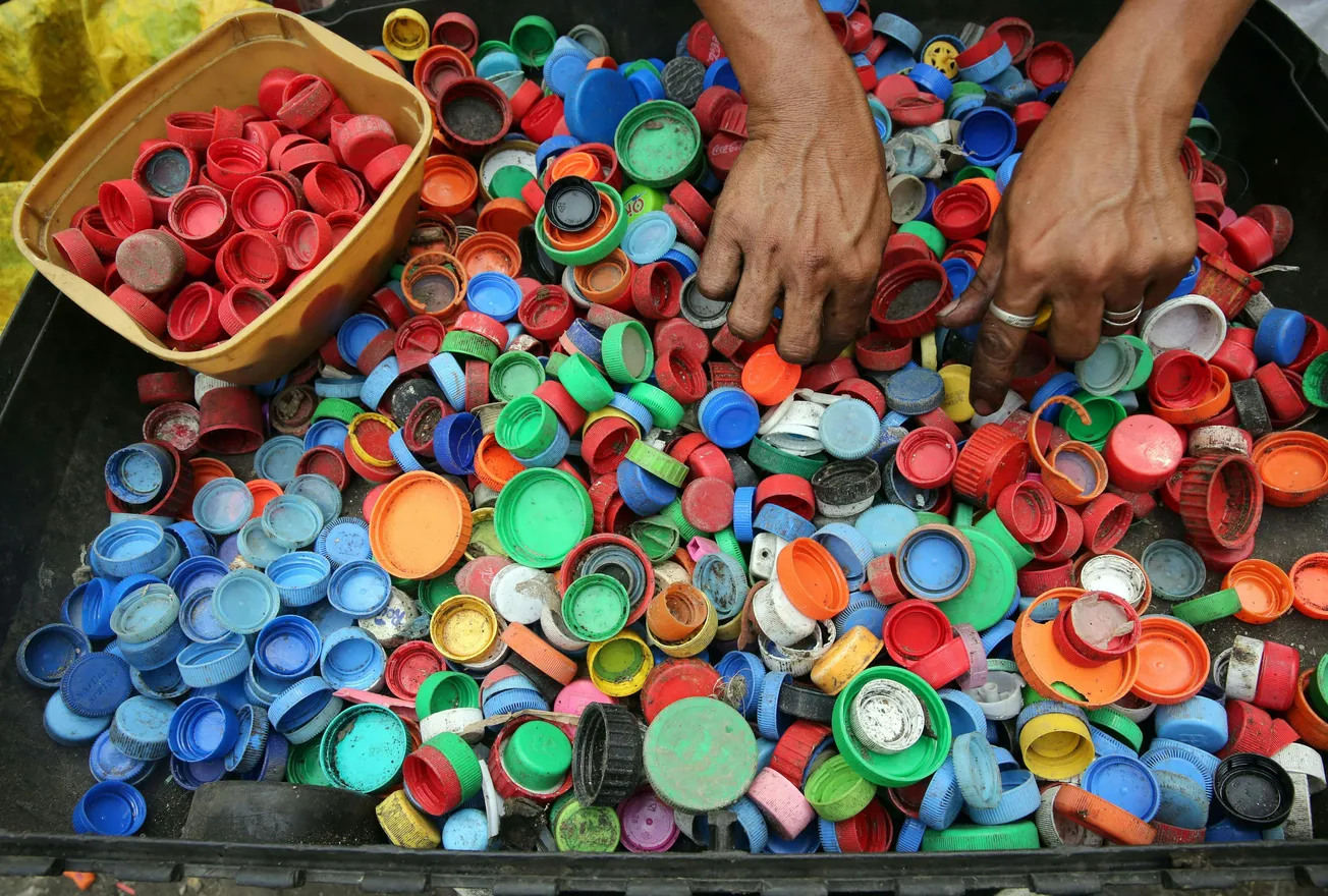Hands sorting colorful plastic bottle caps in a pile, emphasizing recycling. Caps in red, blue, green, and orange; a beige container holds red caps.