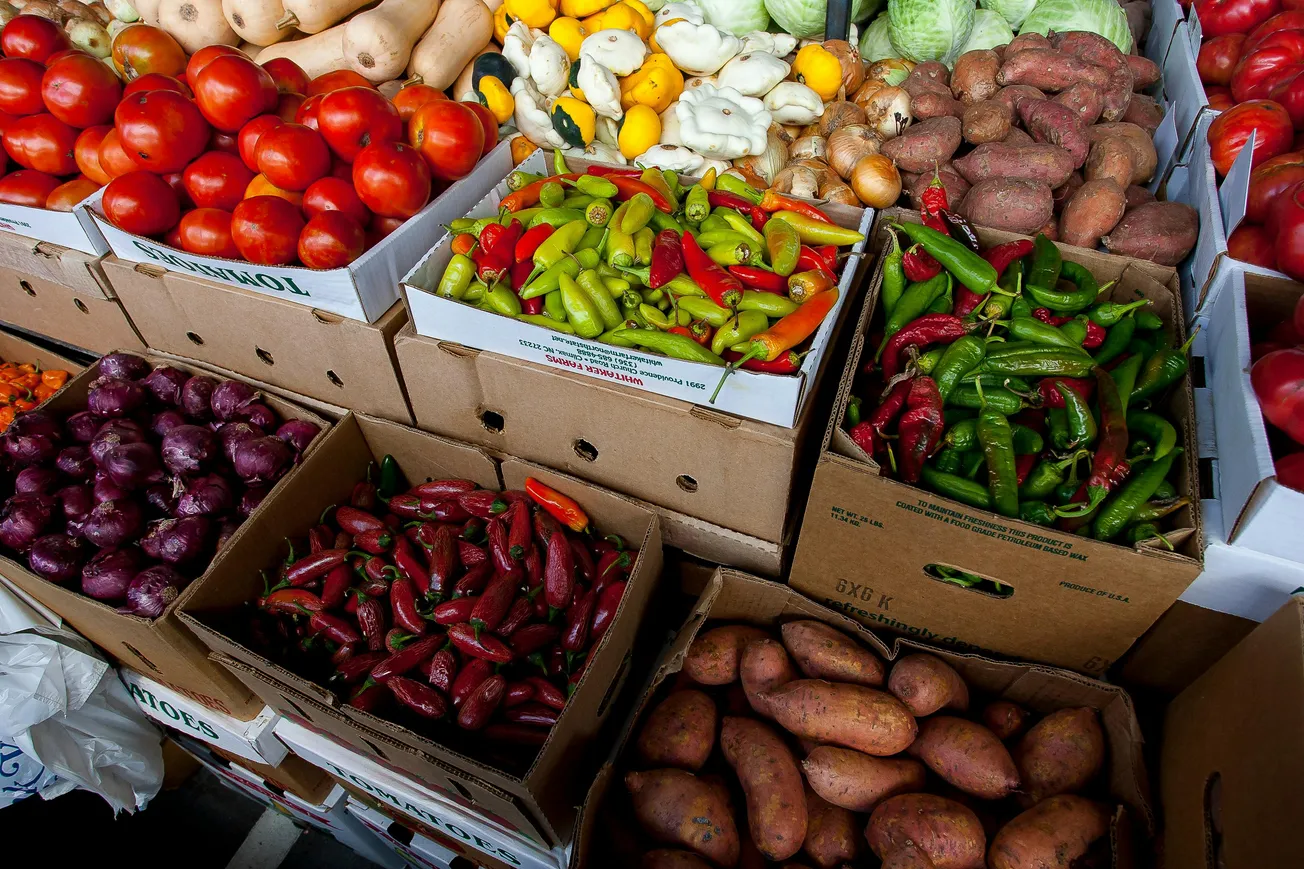Assorted vegetables, including tomatoes, peppers, sweet potatoes, and onions, displayed in cardboard boxes at a vibrant farmers market.