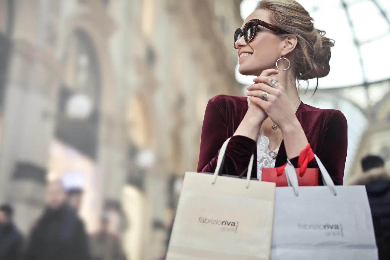 A smiling woman in a red velvet top and sunglasses holds shopping bags in an elegant indoor mall.