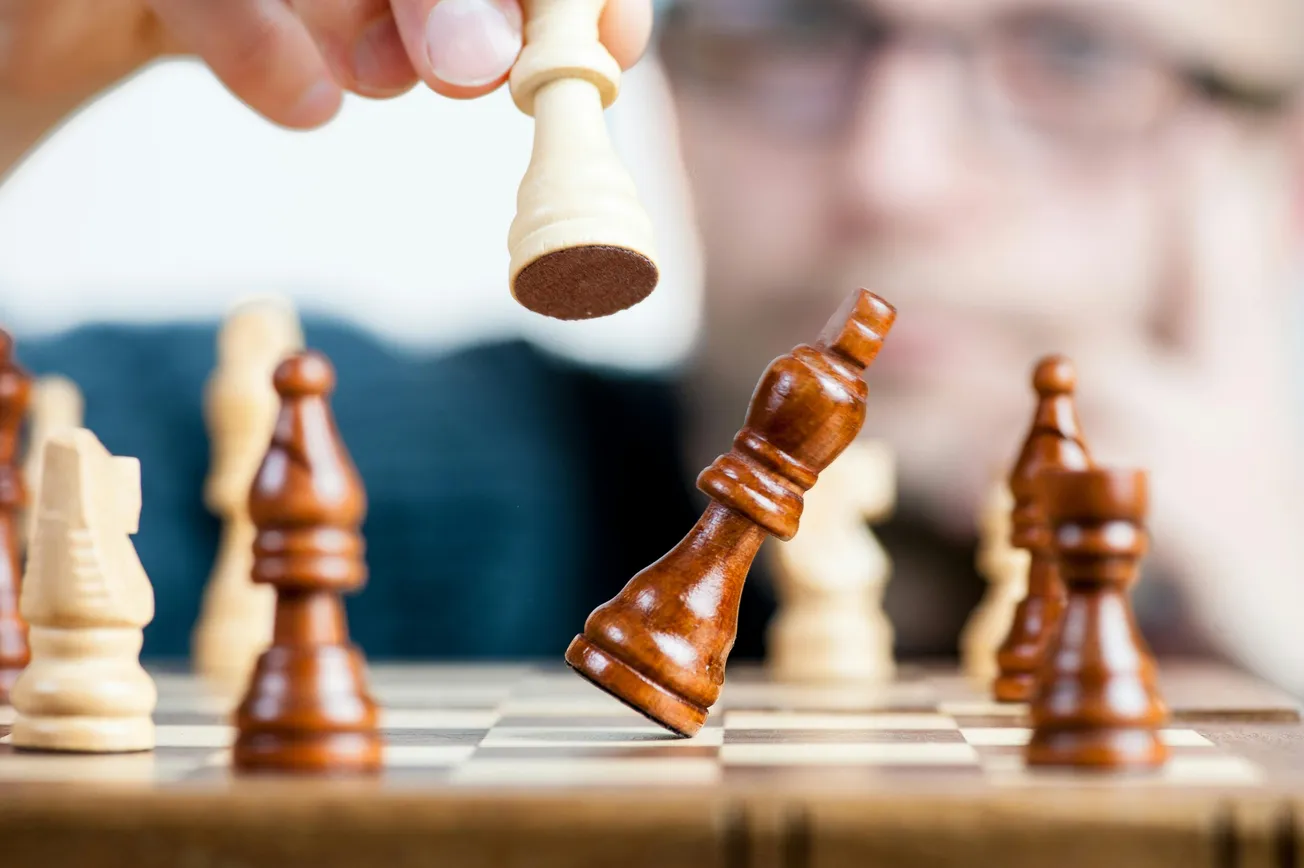 A close-up of a chess game shows a player's hand moving a light-colored piece, knocking over a dark-colored king, suggesting a winning move.