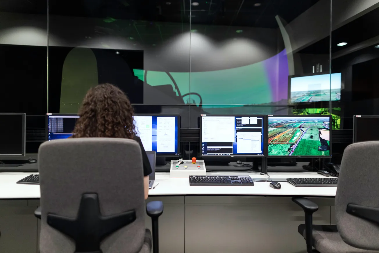 A person with curly hair sits at a control desk in front of multiple monitors displaying software and aerial images, in a control room.