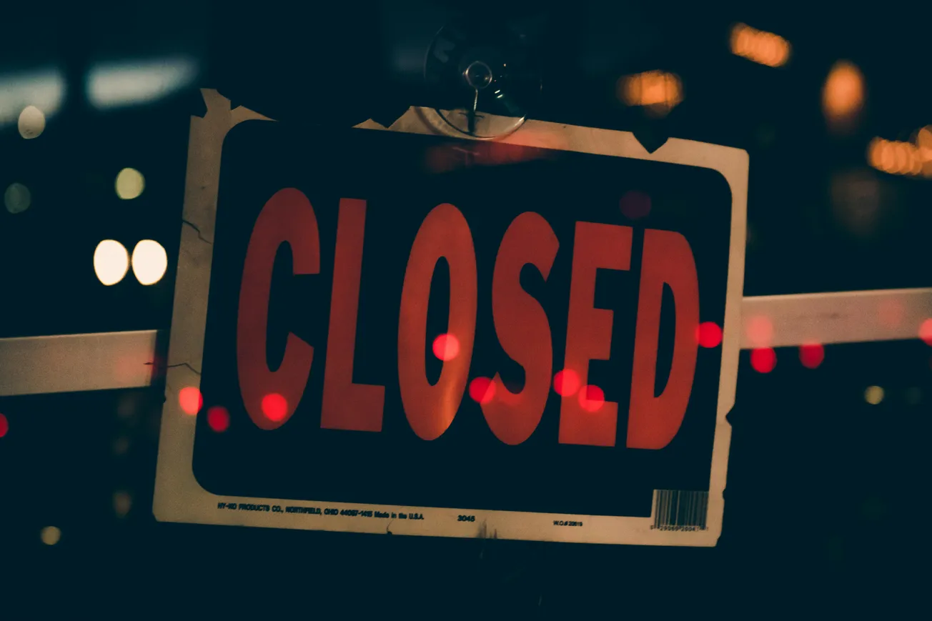 A dark, moody photo of a sign with "CLOSED" in bold red letters, illuminated by out-of-focus red and white lights.