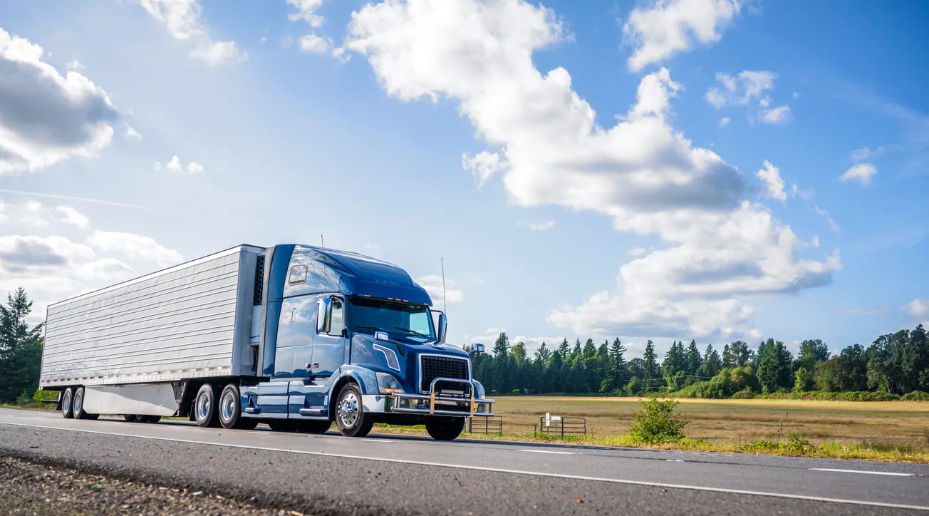 A sleek blue semi-truck drives on a rural road under a bright blue sky with fluffy clouds. Lush green fields and trees line the horizon.