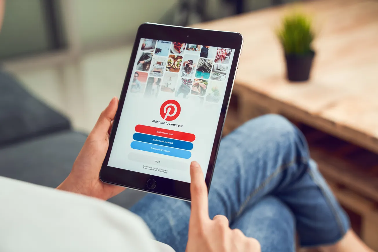 Person holding a tablet displaying the Pinterest login screen, surrounded by colorful pins.