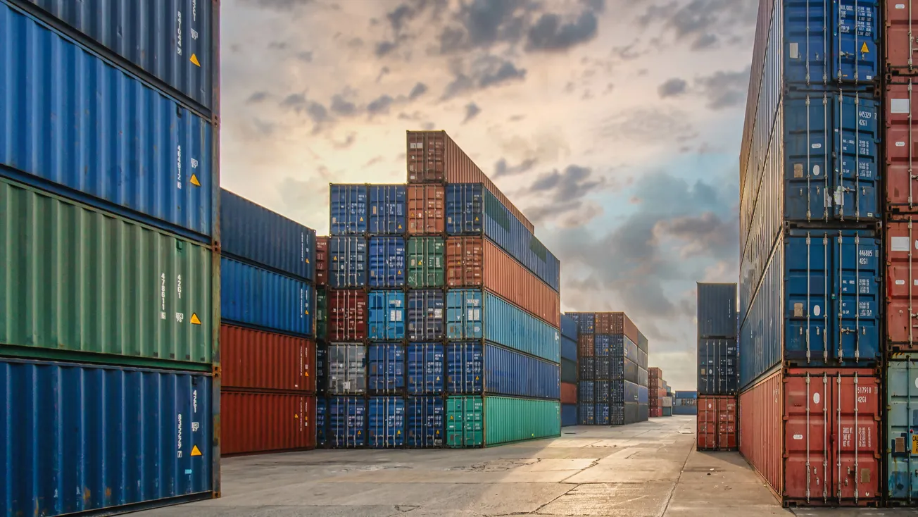 Stacks of colorful shipping containers in a port at sunset. The sky is partly cloudy.