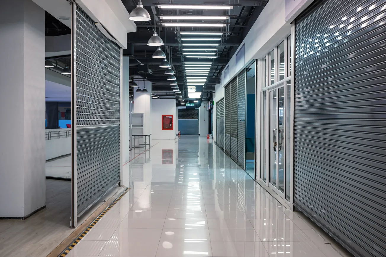 Empty shopping mall corridor with closed metal shutters on both sides. The polished floor reflects fluorescent ceiling lights, creating a serene ambiance.