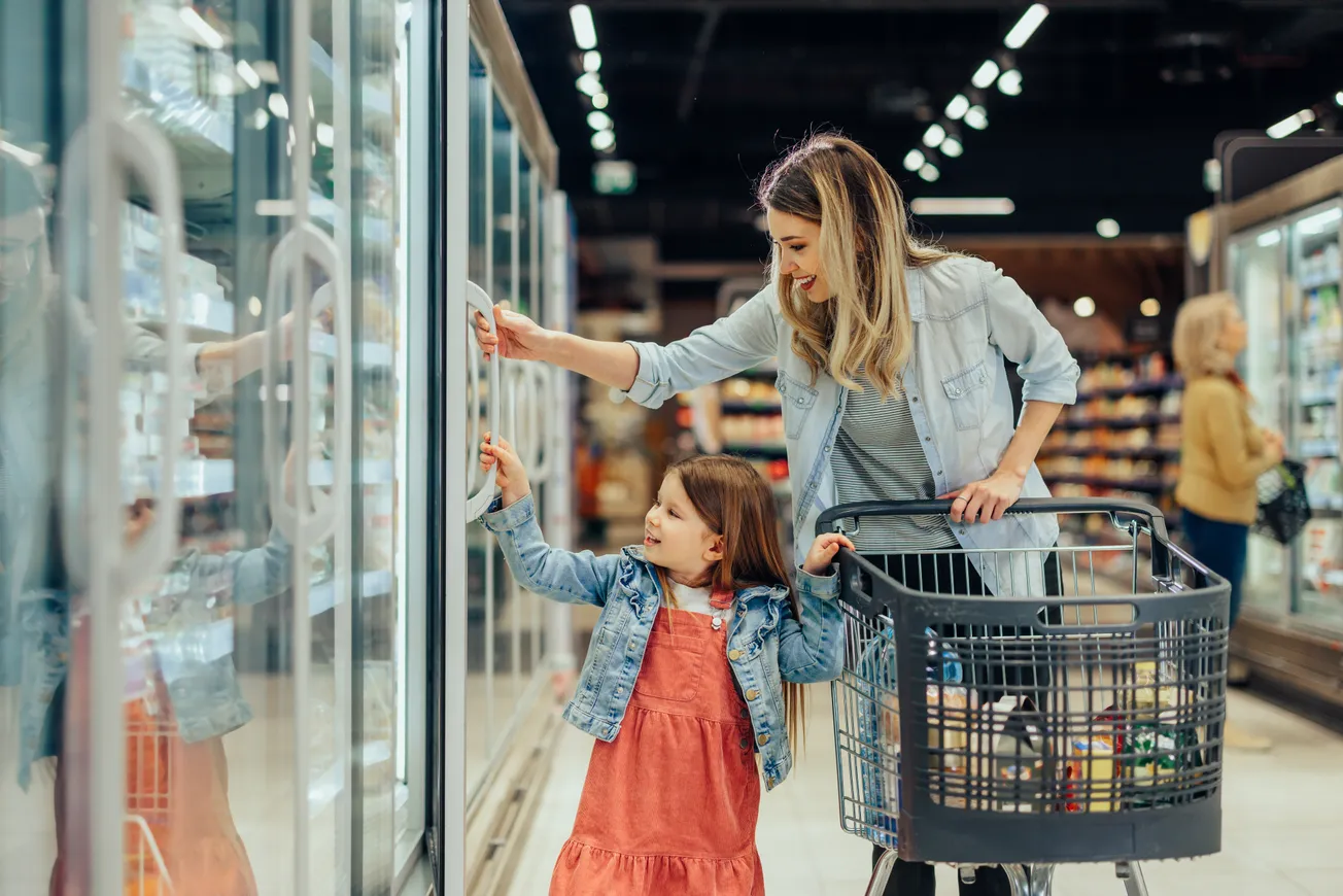A woman and a young girl smile while selecting items from a grocery store freezer. The woman pushes a shopping cart, creating a warm, family-oriented scene.