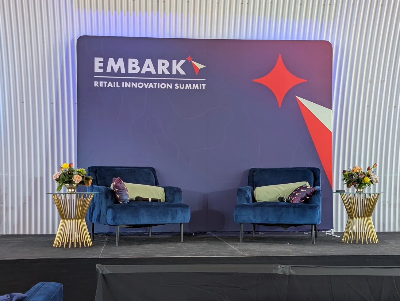 Stage setup at the Embark Retail Innovation Summit with two blue armchairs and gold side tables against a branded backdrop.