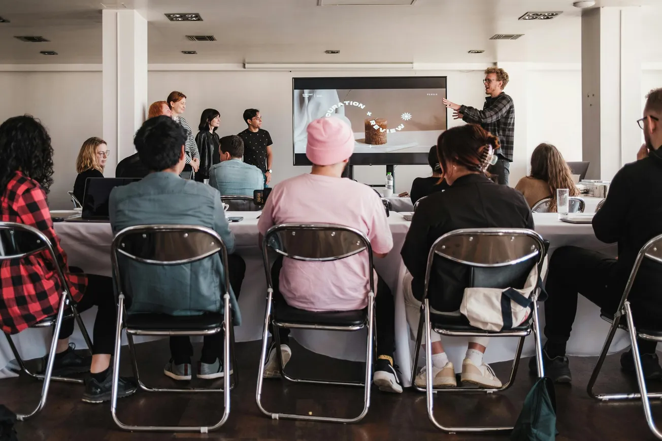 A group of people in a classroom setting listens to a presentation. A speaker gestures towards a screen. Attendees sit at a table, creating an engaged atmosphere.