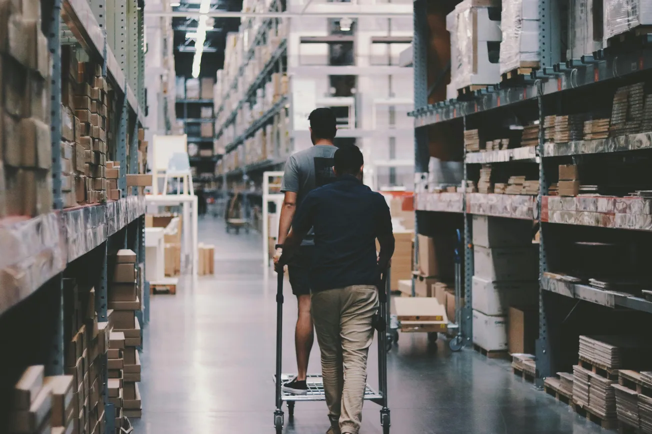 Two people in a warehouse with high shelves filled with boxes and packages. One is pushing a cart while the other rides on it, suggesting teamwork.