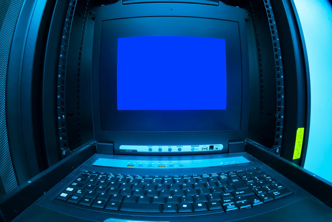 A server rack with a laptop sporting a blank blue screen. The room is dimly lit, creating a technical, focused atmosphere.