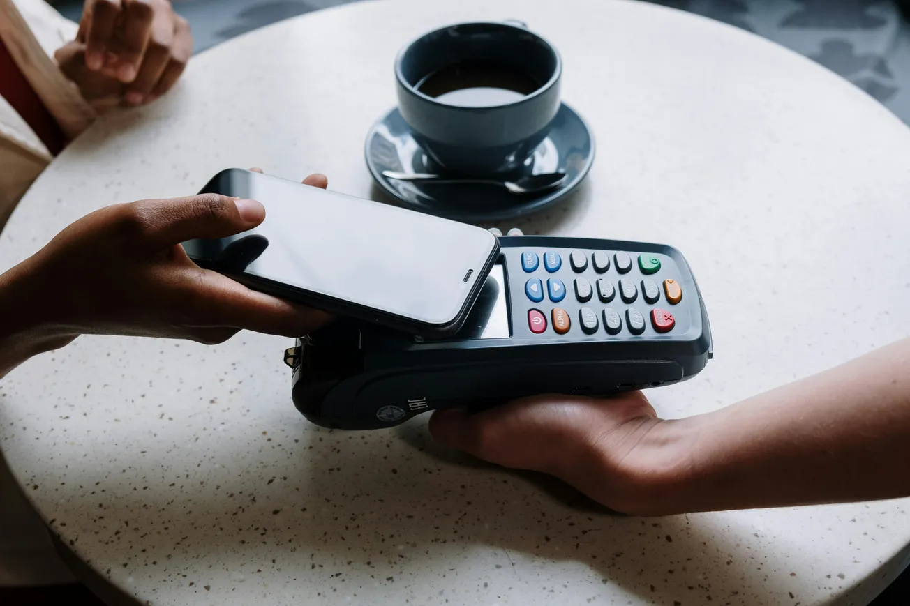 A smartphone is held over a payment terminal for contactless payment on a round table. A coffee cup is in the background.