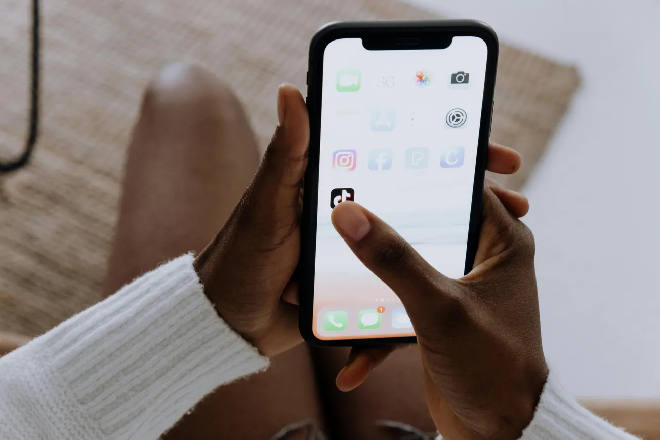 Two hands holding a smartphone displaying app icons on a screen, including social media apps, against a blurred background of legs and a beige rug.