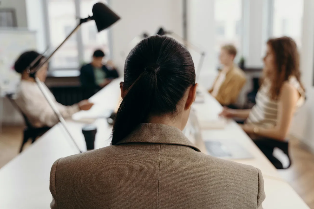 A group of people sits around a conference table in a bright office. Focus is on a person with a ponytail in the foreground. The atmosphere is professional.
