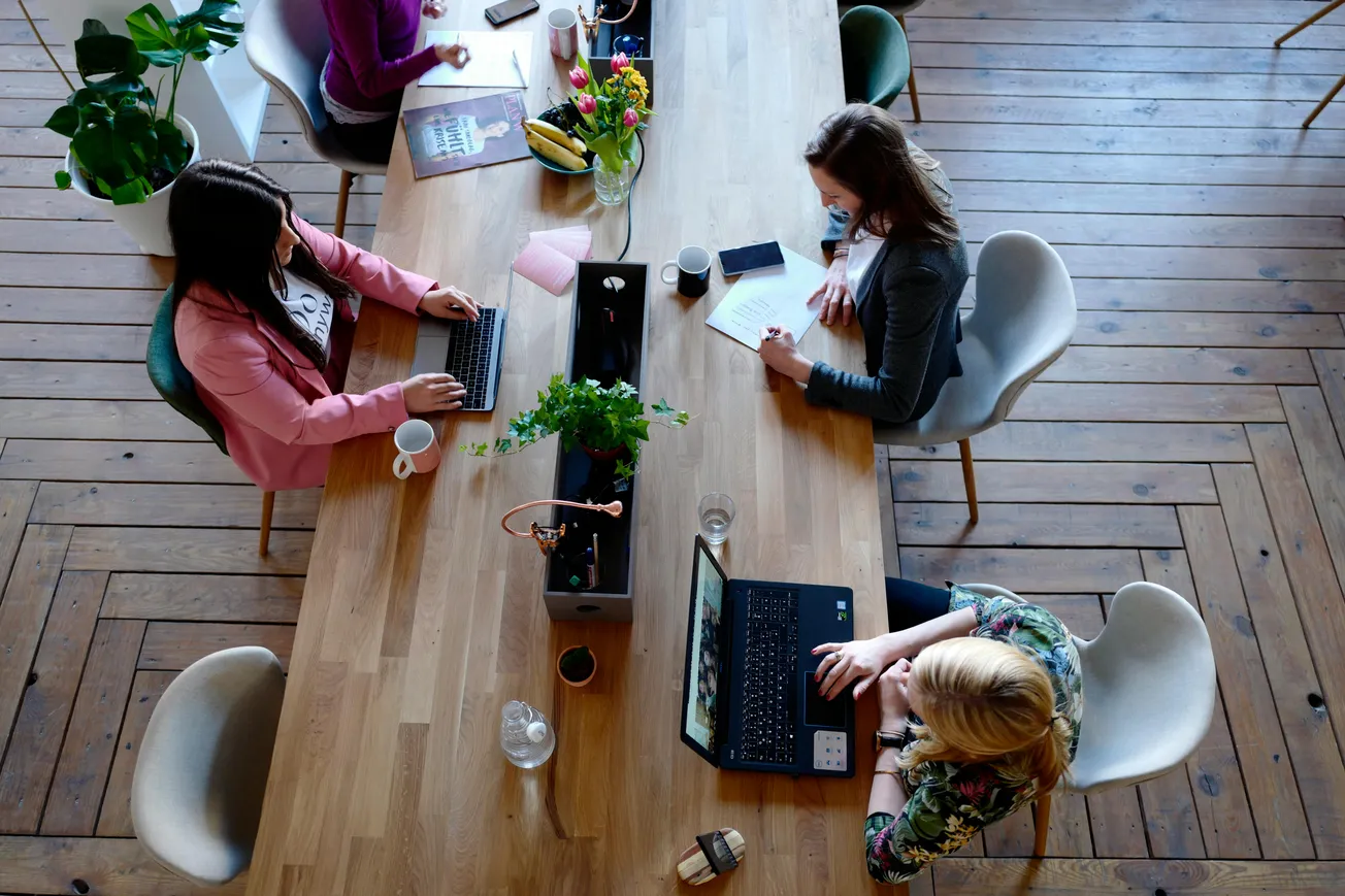 Overhead view of a shared wooden desk with three people working on laptops and writing. Bright, modern space with plants.