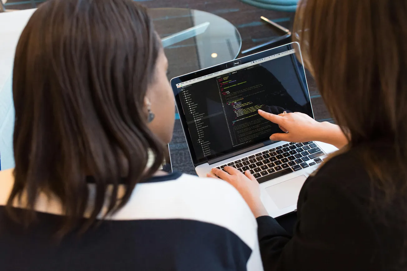 Two women sit at a desk, focused on a laptop displaying code. One woman points at the screen.
