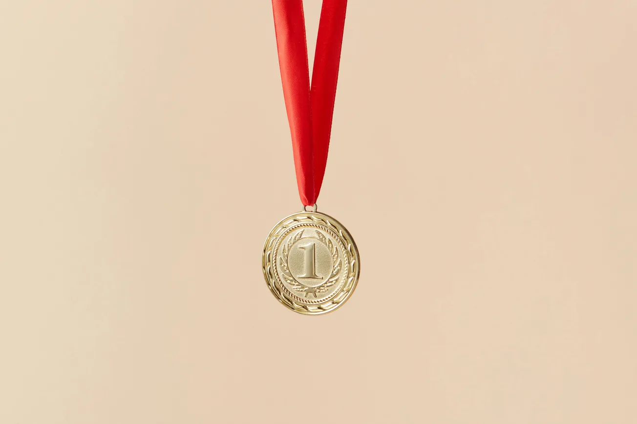 Gold medal with a number "1" embossed, hanging from a red ribbon on a light beige background, conveying achievement and victory.