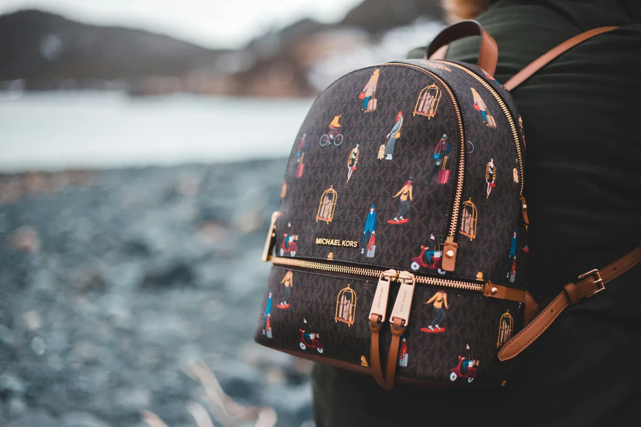 A person wearing a dark backpack, featuring colorful illustrated patterns, stands near a rocky shoreline. The backpack conveys a stylish, adventurous vibe.