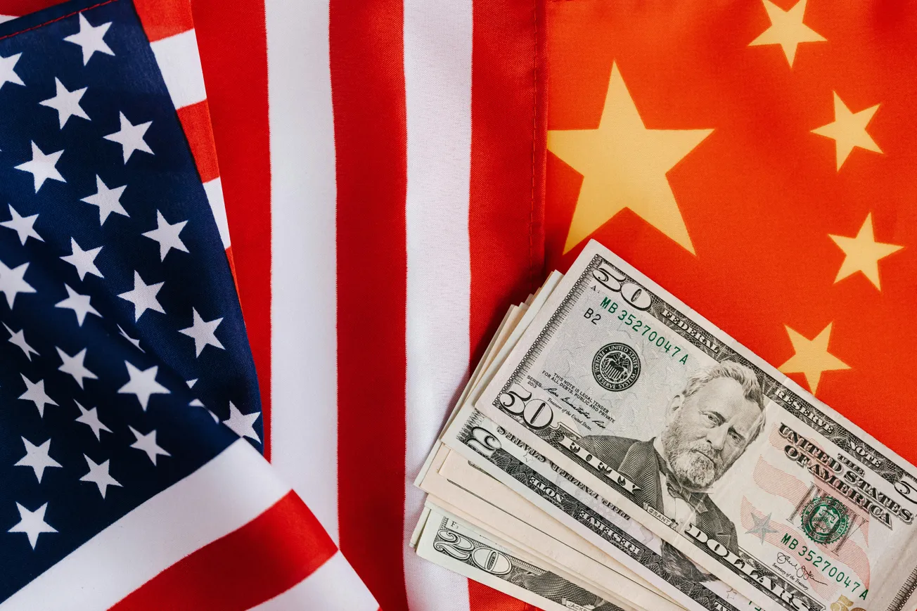 Two overlapping flags, one American and one Chinese, with a stack of U.S. dollar bills on the Chinese flag. The image conveys international finance.