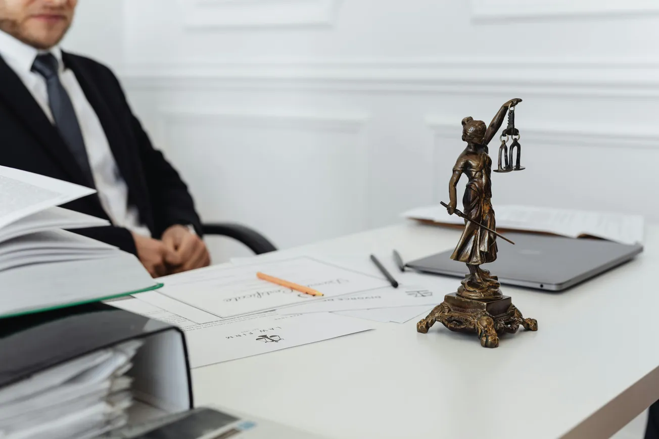 A person in a suit sits at a desk with legal documents. A bronze Lady Justice figurine is on the desk, conveying a serious and professional tone.