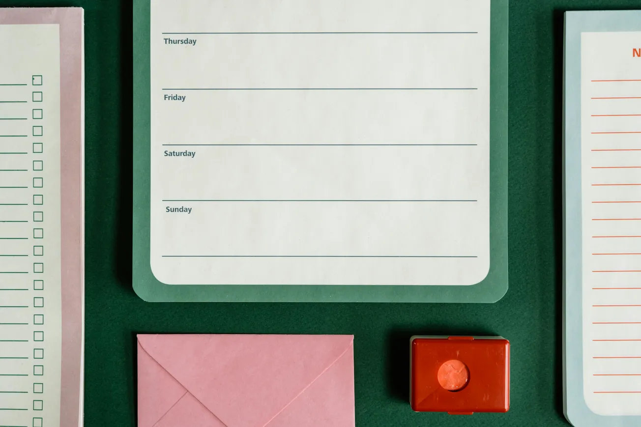A green surface with a weekly planner, checklist, and lined paper. A pink envelope and red stamp pad add a touch of color.