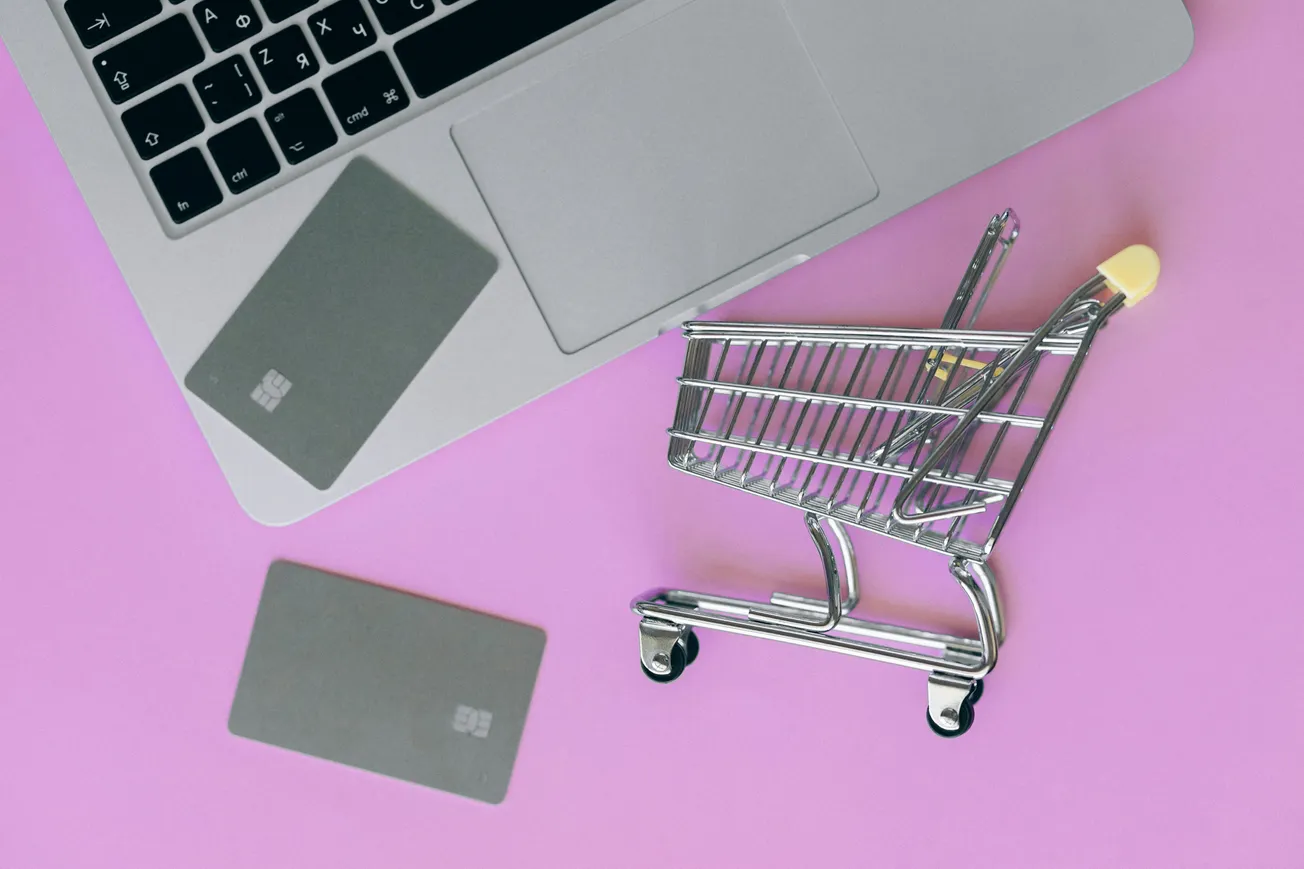 Laptop and credit cards on a pink surface with a small shopping cart nearby, symbolizing online shopping. The scene conveys a modern, digital commerce vibe.