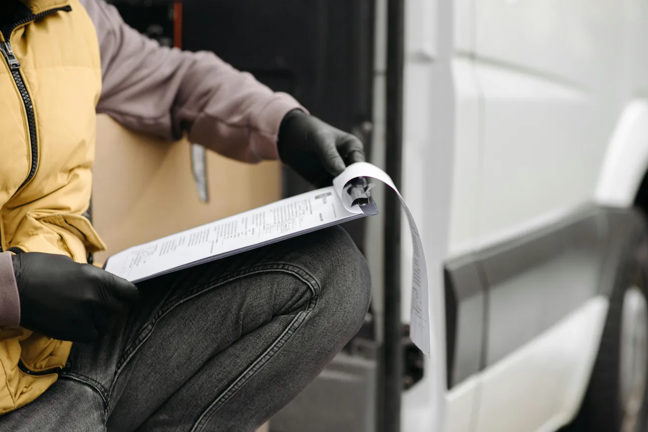 A person wearing a yellow vest and black gloves holds a clipboard with papers, sitting on the edge of a van.