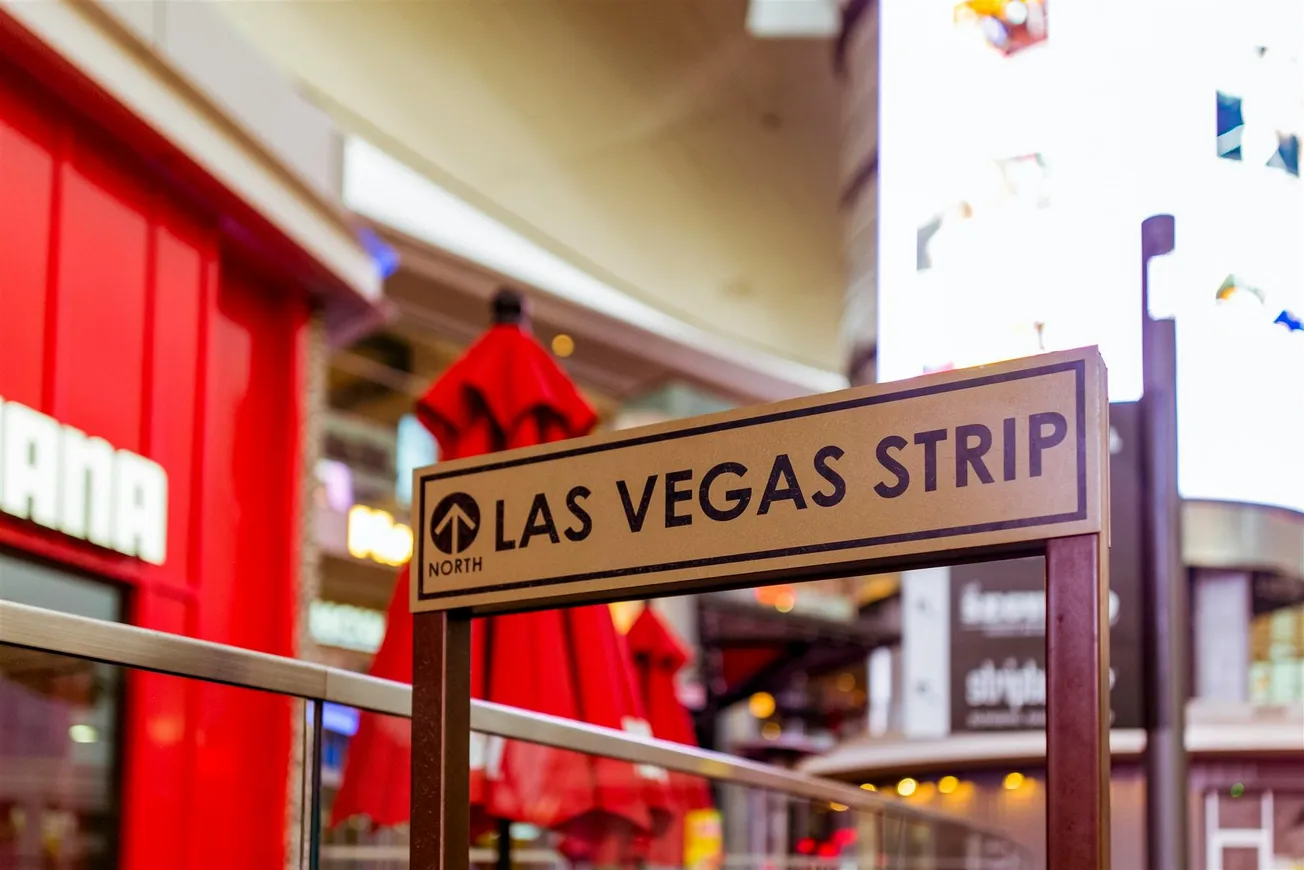 Sign reading "Las Vegas Strip" in a bustling scene with bright red umbrellas and illuminated buildings, conveying a lively and vibrant atmosphere.