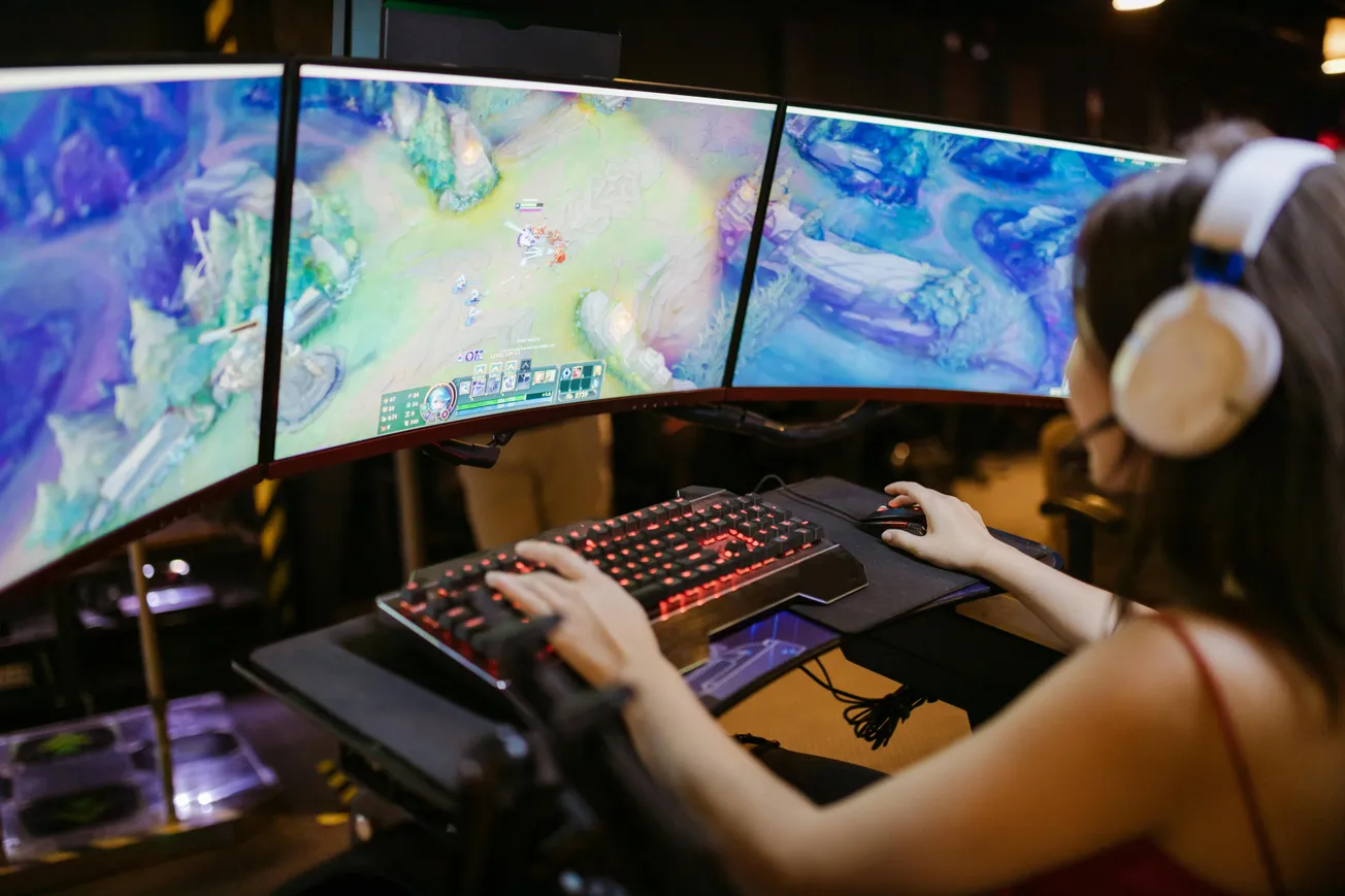 A gamer with headphones is focused on playing on a setup with three curved monitors displaying a vibrant game. A glowing keyboard adds to the immersive experience.