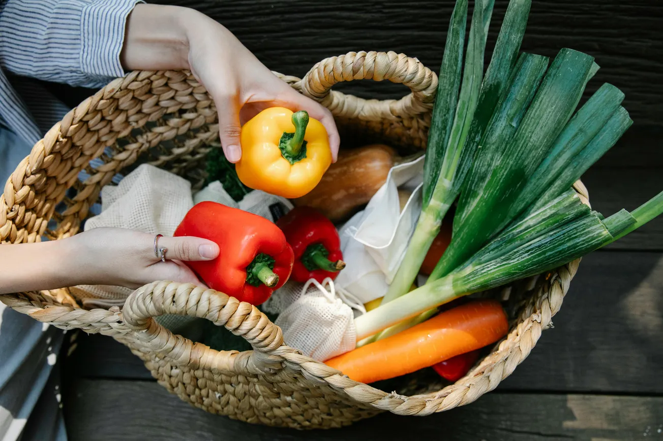 A woven basket holds assorted fresh vegetables, including leeks, carrots, and bell peppers. Two hands pick a yellow pepper.