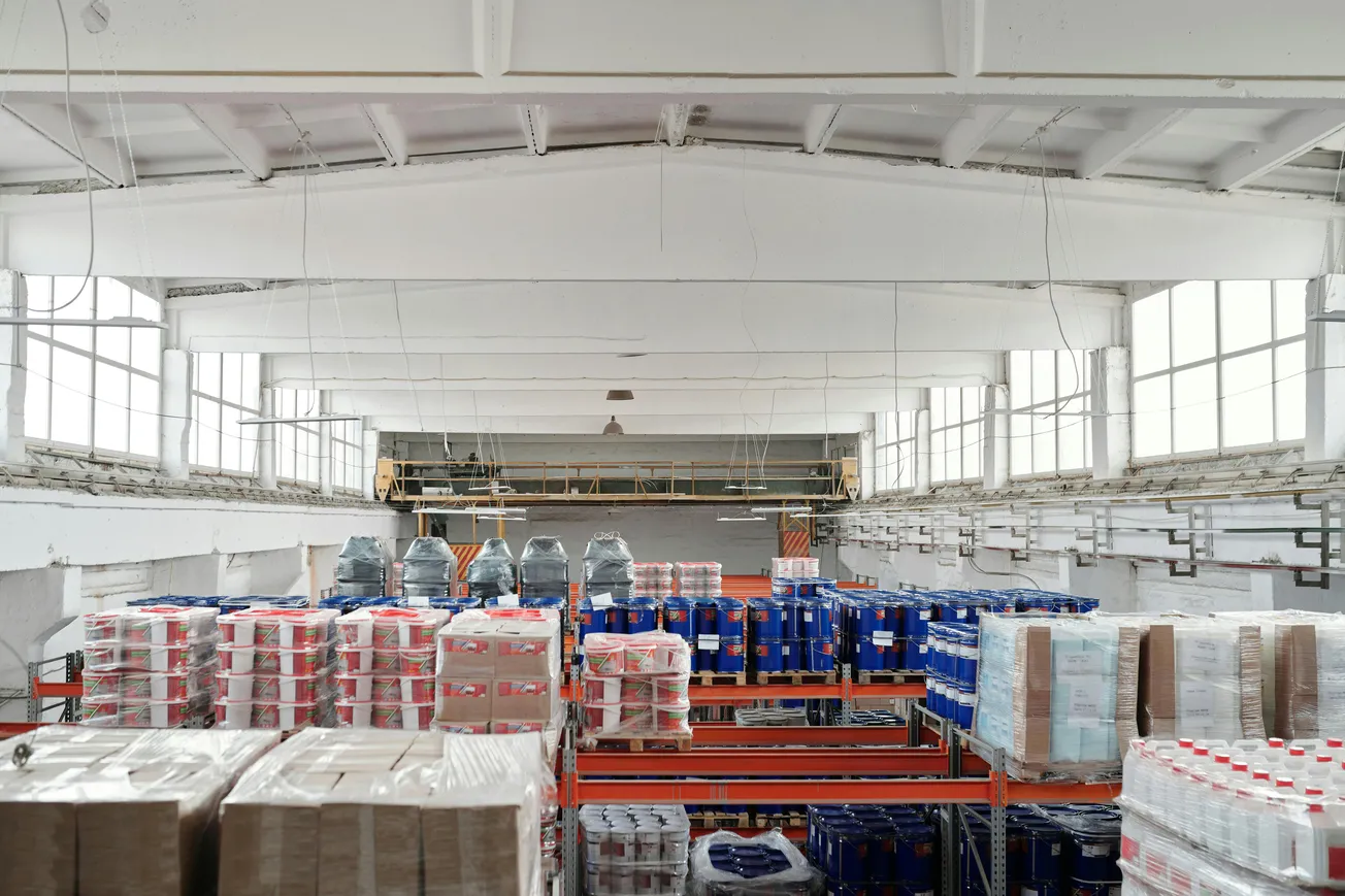 A spacious industrial warehouse with high ceilings, filled with stacked pallets of goods in plastic wrapping. Natural light streams through large windows.