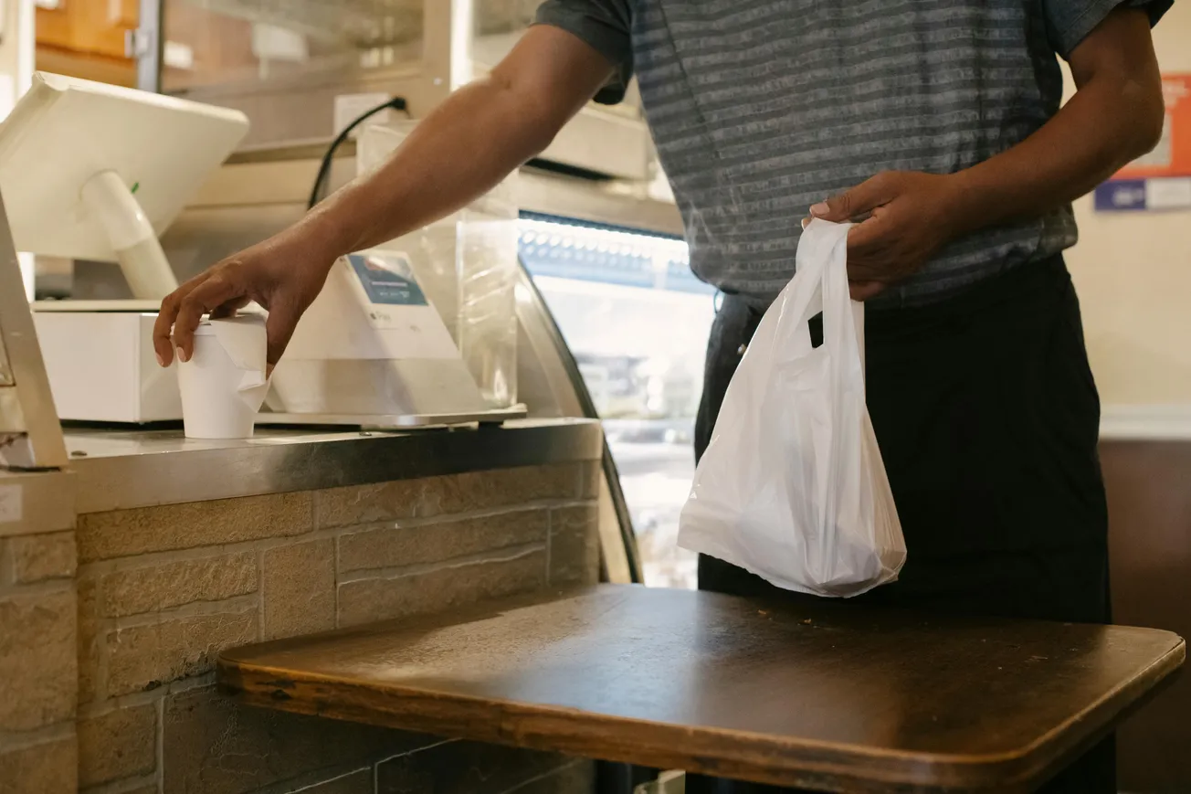 A person in a striped shirt places a takeout coffee cup on a counter, while holding a white plastic bag. The setting is a warm café environment.