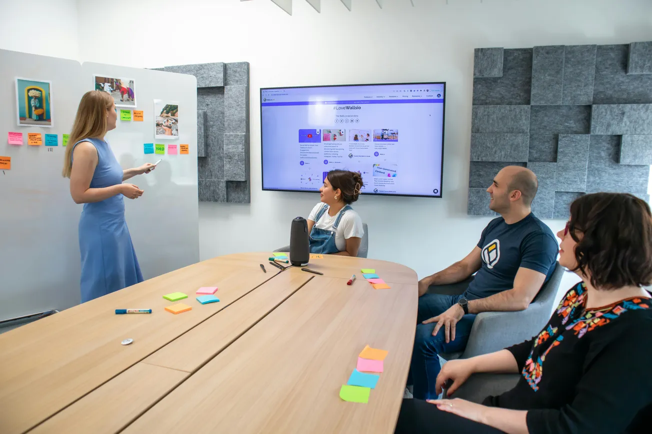 A woman in a blue dress presents at a whiteboard covered in colorful sticky notes, while three people seated at a table listen attentively. A screen displays a website.