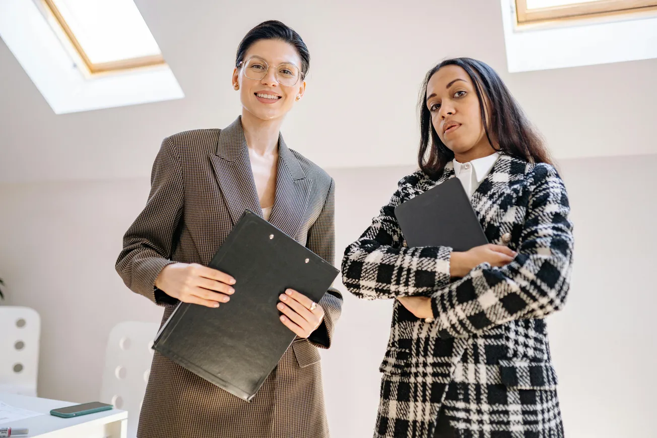 Two confident businesswomen in stylish suits hold black folders, standing in a bright office space. They convey a professional and assertive tone.