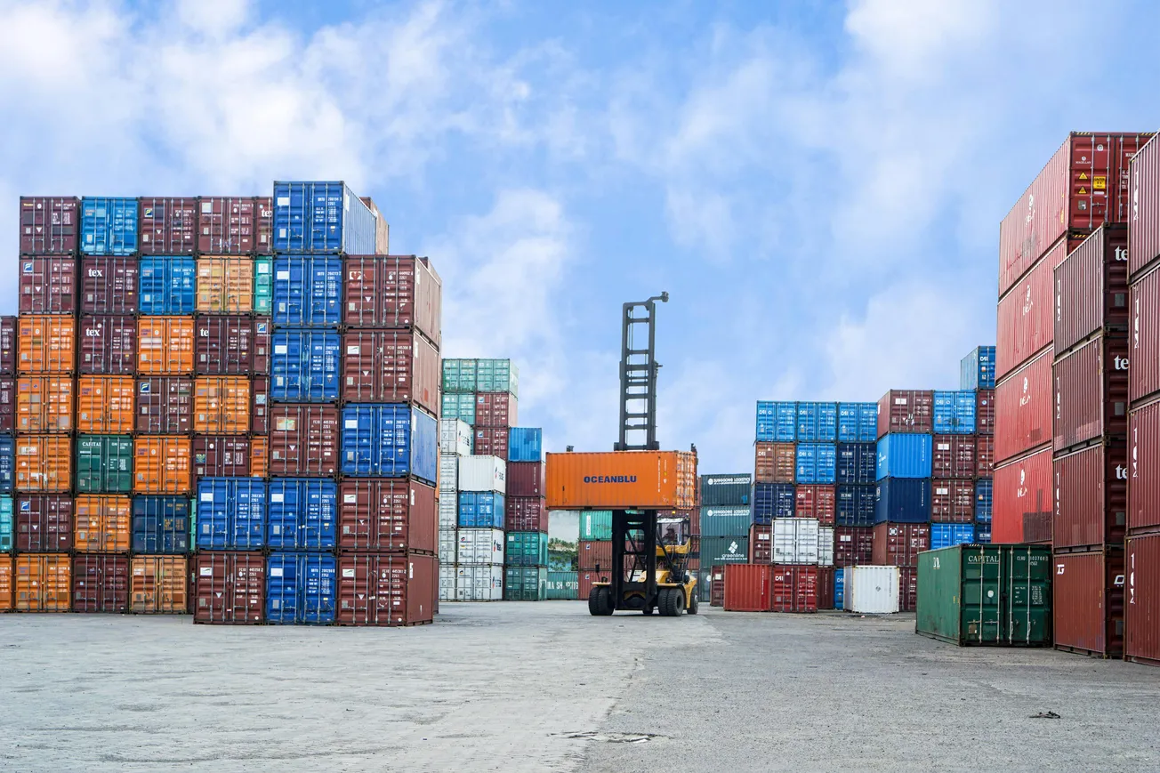 A forklift moves a bright orange shipping container in a port yard, surrounded by colorful stacks of containers under a blue sky with clouds.