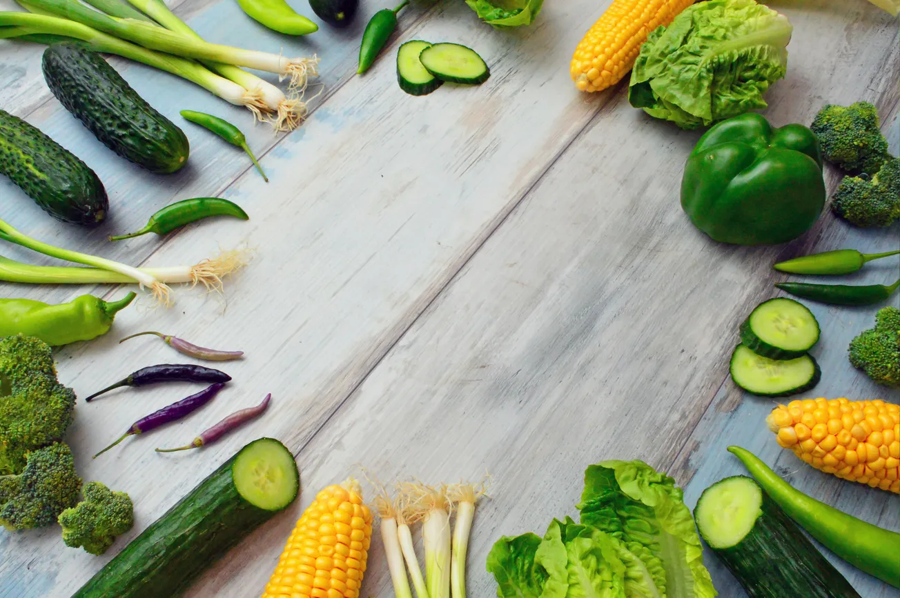 A variety of fresh vegetables are arranged in a semicircle on a light wooden surface. Veggies include cucumbers, green peppers, corn, lettuce, and green onions.