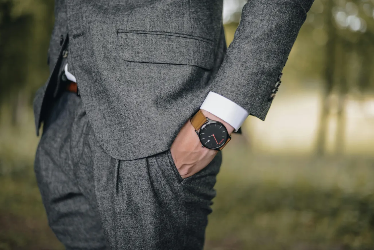 A person wearing a tailored gray suit with a brown leather watch, standing outdoors. The left hand is in the pocket, conveying a sense of elegance.
