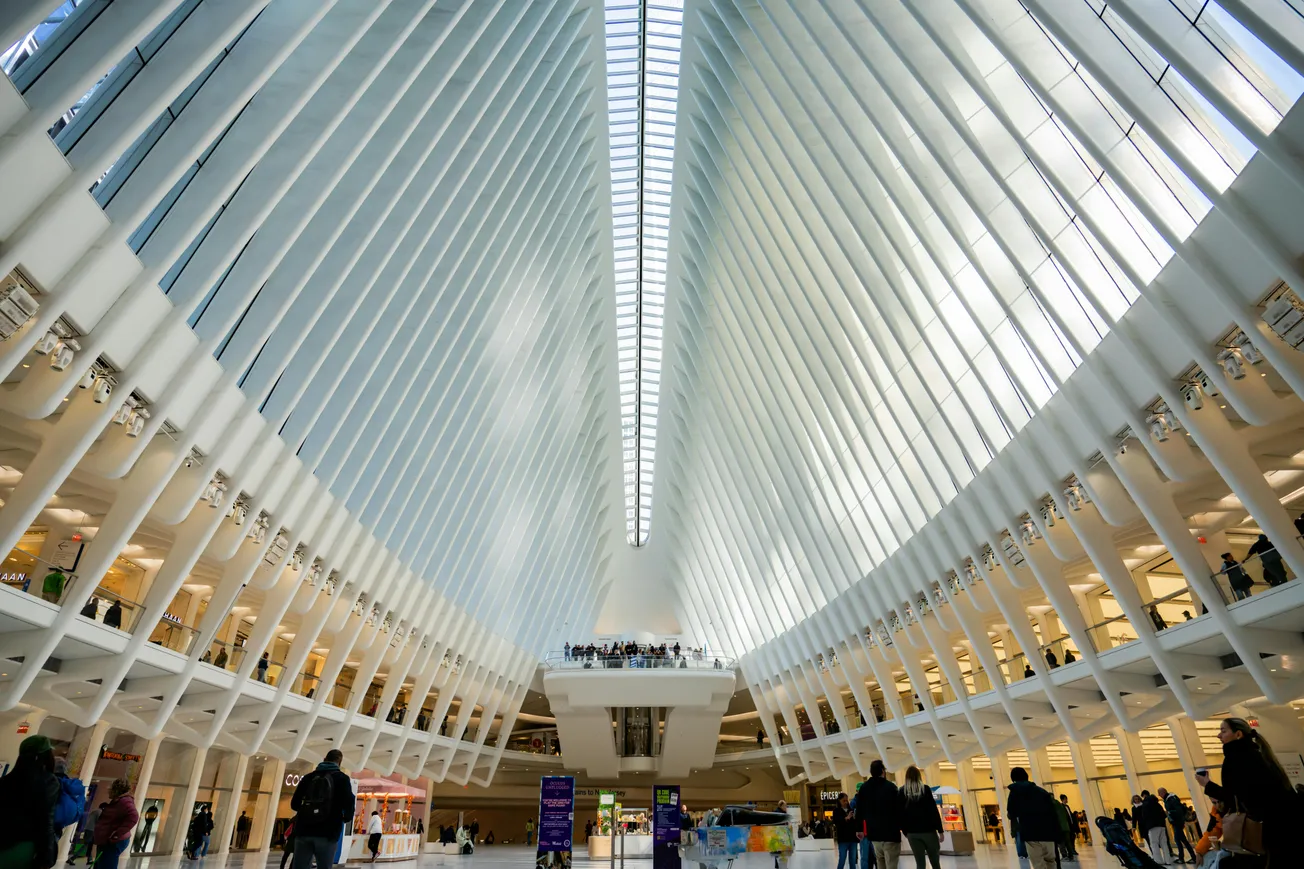Futuristic interior of a transit hub with soaring white ribbed arches and large skylight, bustling with people. The space feels open and dynamic.