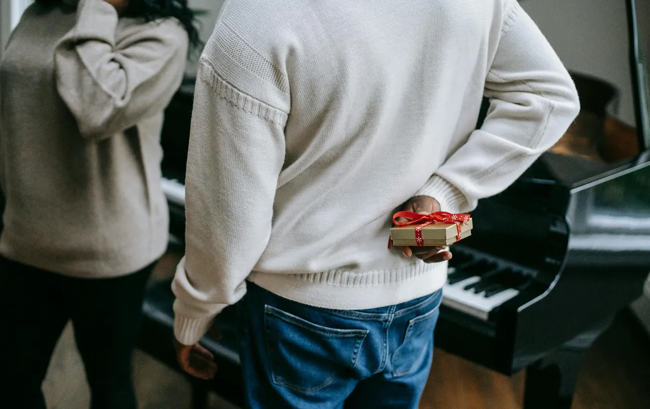 A person in a cream sweater and jeans hides a small gift wrapped in red ribbon behind their back, standing near a piano, conveying a sense of surprise.