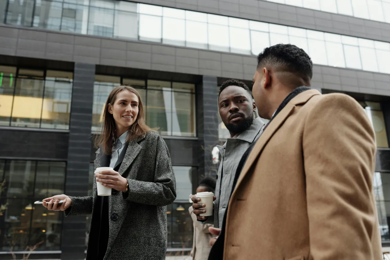 Three people in coats walking and chatting outdoors, each holding a coffee cup. They seem engaged and happy, with a modern glass building behind them.